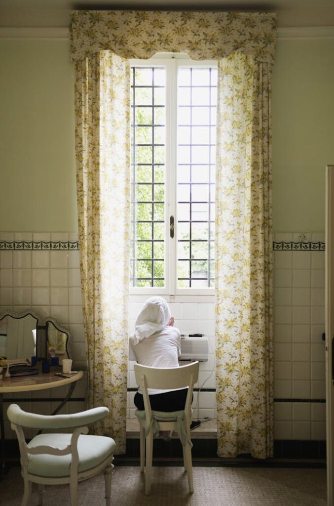 A woman in a cozy room with floral curtains gazing out a window, creating a peaceful ambiance.